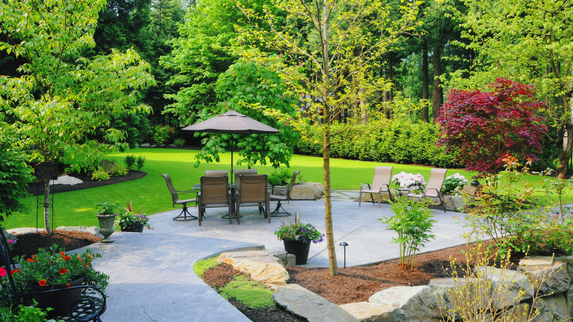 Landscaped backyard with stone patio, outdoor furniture, and lush green trees